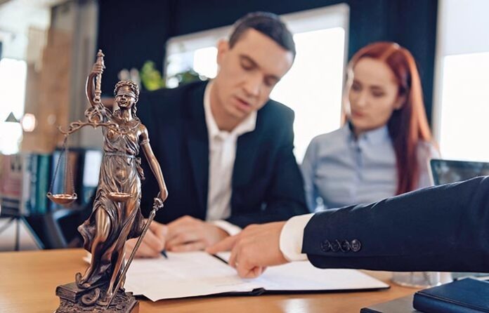 Statue of Themis holds scales of justice. In unfocused background, adult man signs documents.