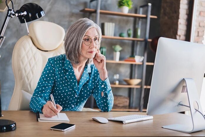 Photo of attractive calm serious grey haired old woman look pc rite note indoors in workplace workstation